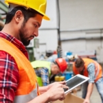Handwerker tippt im Lager stehend auf seinem Tablet - 2 Kollegen arbeiten im Hintergrund